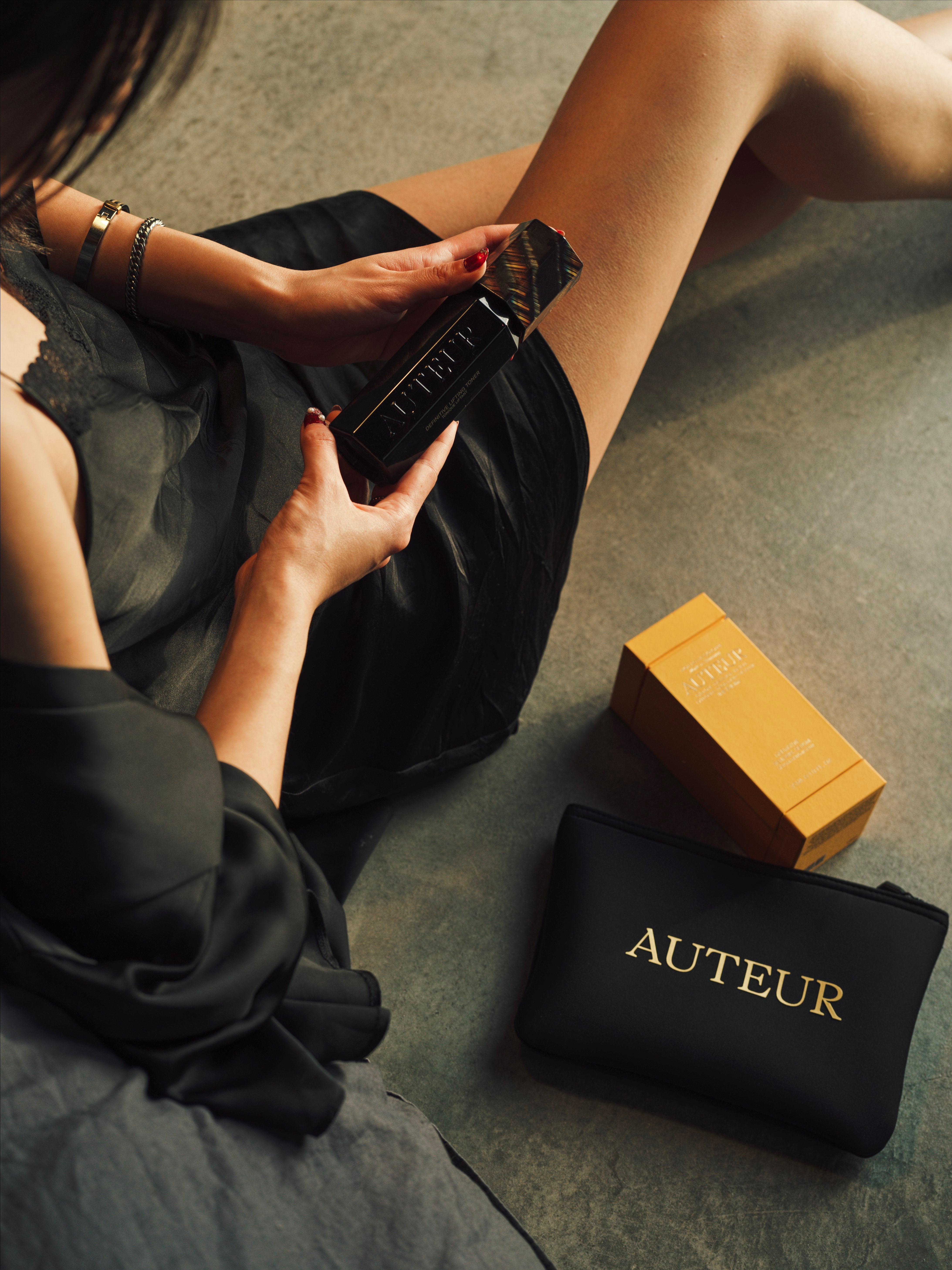 Person holding a black bottle with a gold cap, next to a black pouch labeled 'Auteur' on a concrete floor.