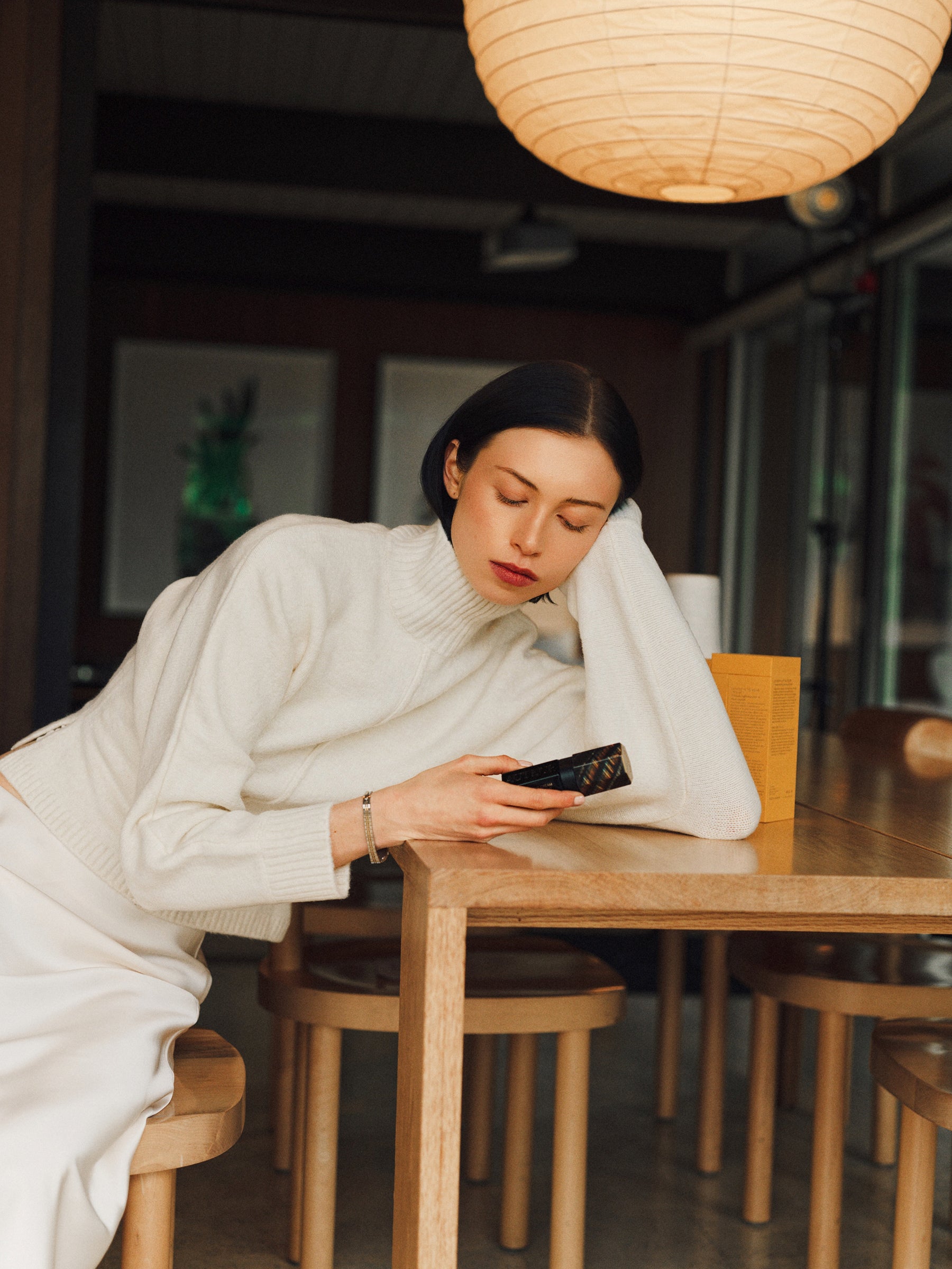 Woman sitting at a wooden table in a dimly lit room, looking at her phone.