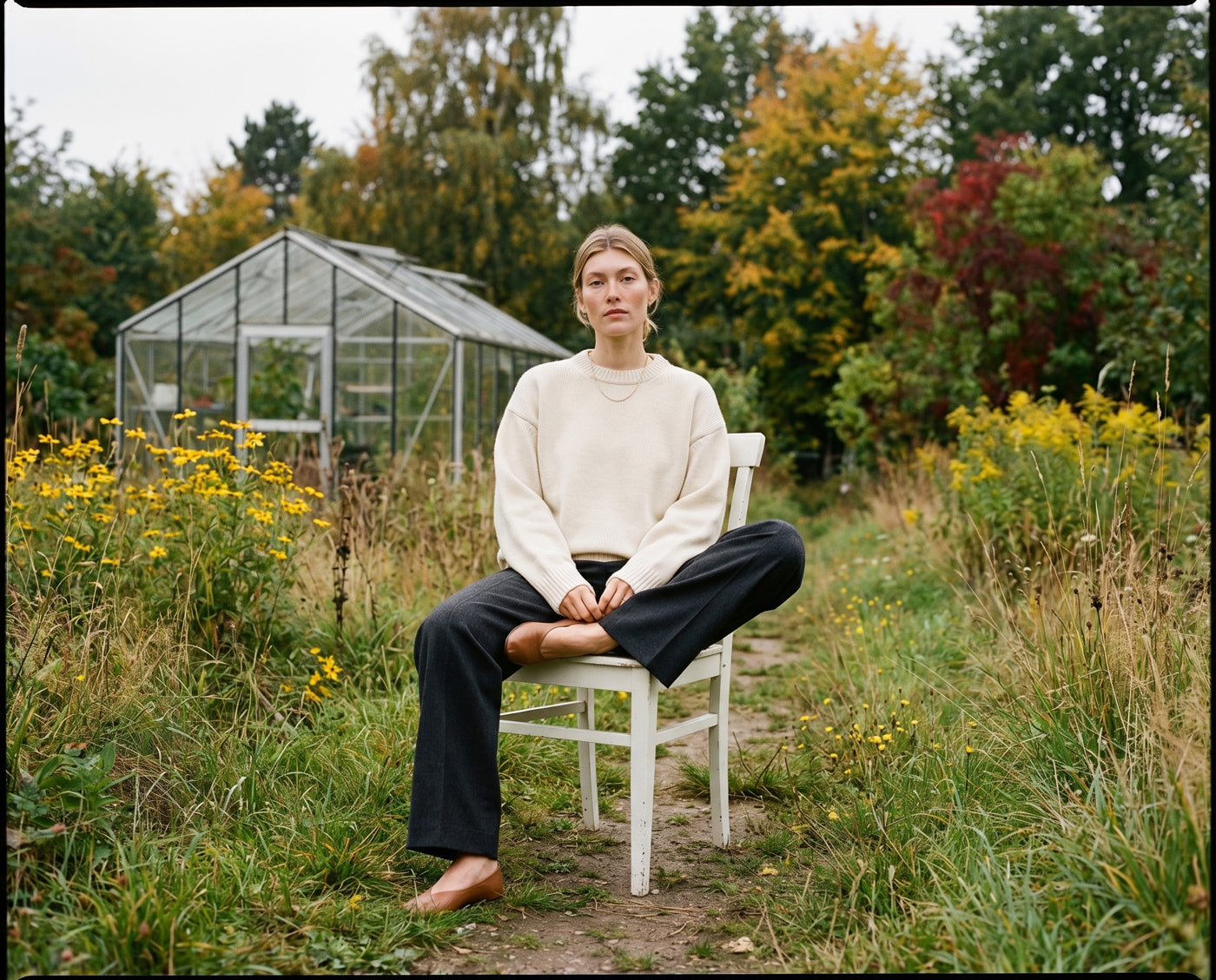 Person sitting on a chair in a garden with greenhouses and trees in the background