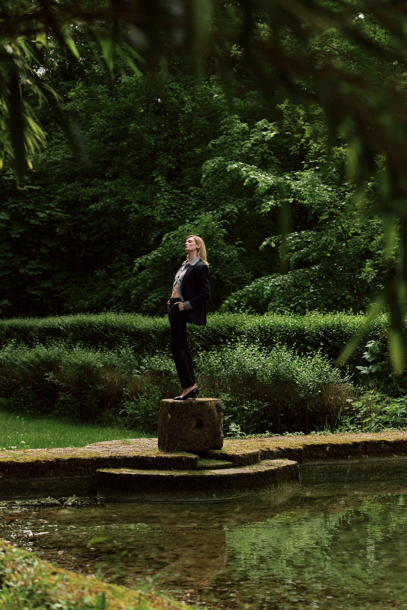Person standing on a stone pedestal by a pond with greenery in the background