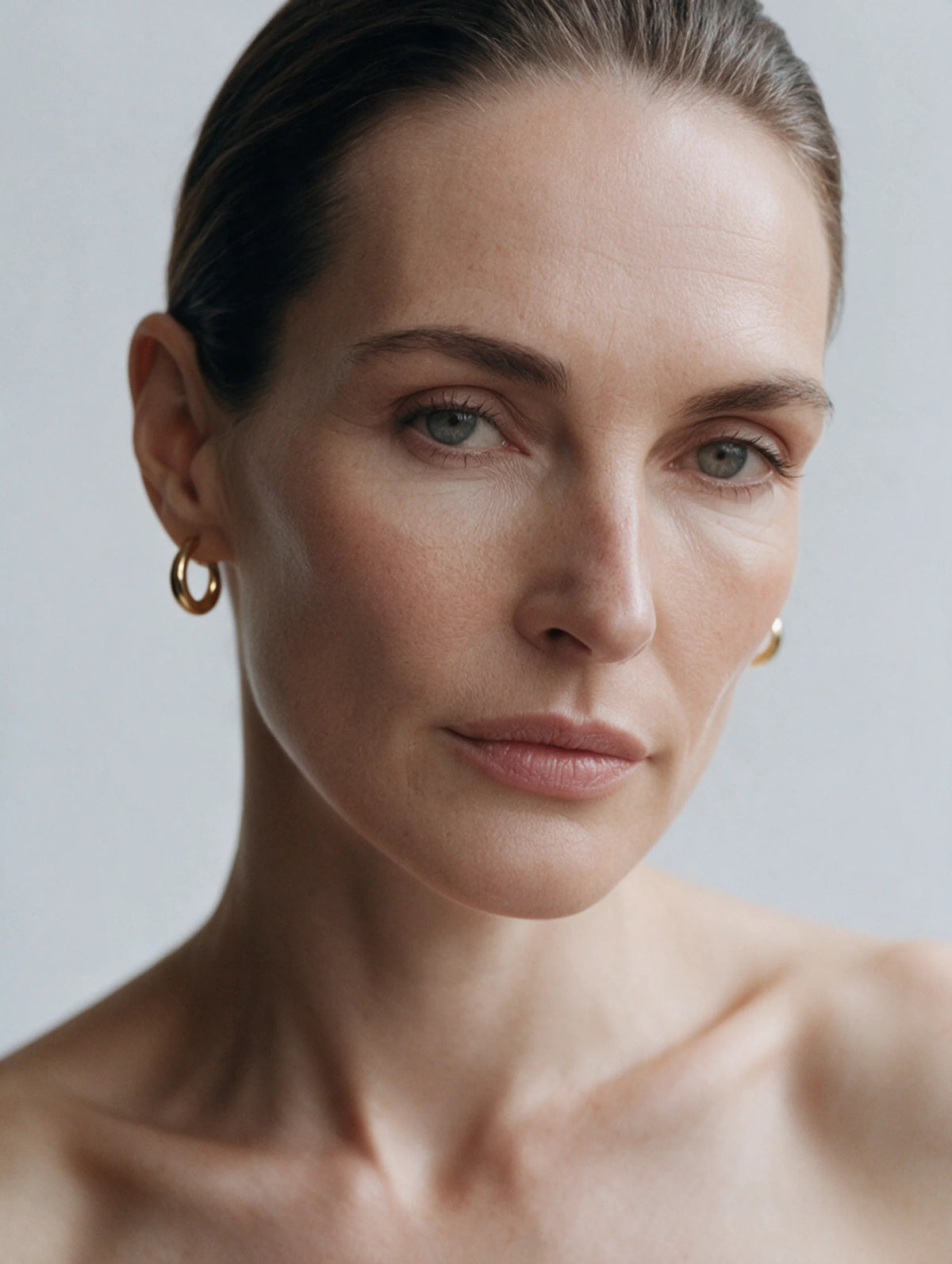 Portrait of a woman with slicked-back hair and gold hoop earrings against a pale grey ground