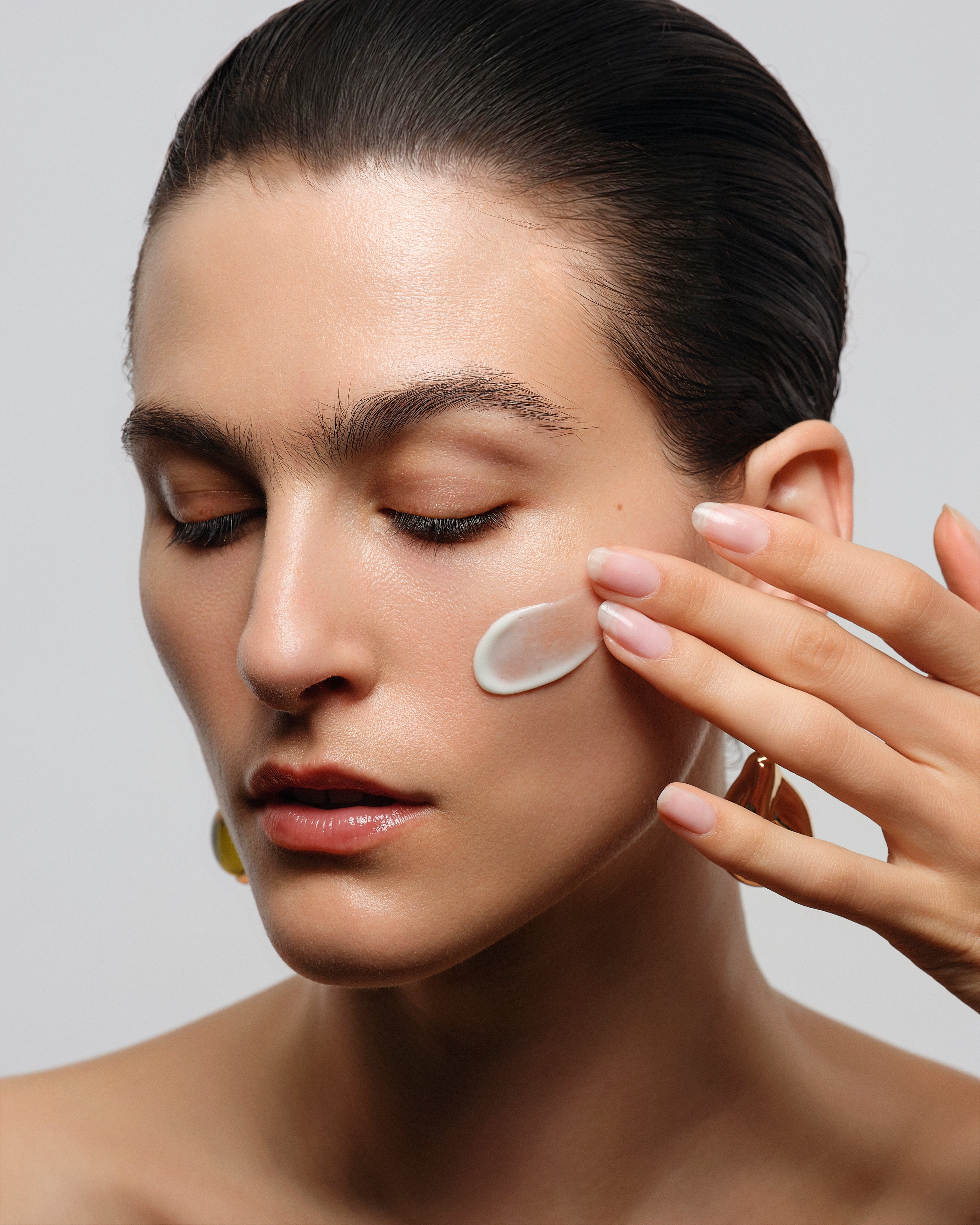 Close-up of a woman with her eyes closed, applying a small white cream or patch to her cheek with her finger, set against a plain light gray background.